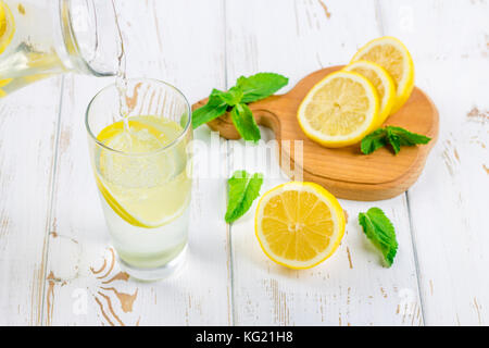 In einem Becherglas, eine kalte Limonade wird aus einer Kanne auf einem weißen Hintergrund Holz umgeben von Zitronen gegossen Stockfoto