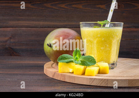 Smoothies Mango mit Stroh und Minze in ein Becherglas. Platte mit in Scheiben geschnittenen Mango. Eine tropische Frucht. Holz- Hintergrund Stockfoto