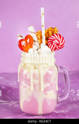 Freakshake von rosa Smoothie, Creme. monstershake mit Lutscher, Waffeln und Eibisch. extreme Milchshake in einem Marmeladenglas. rosa lila Hintergrund. Stockfoto