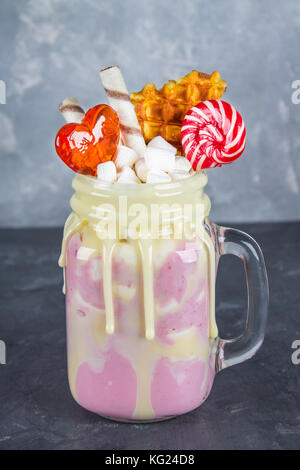 Freakshake von rosa Smoothie, Creme. monstershake mit Lutscher, Waffeln und Eibisch. extreme Milchshake in einem Marmeladenglas. grau dunkel konkrete Hinterg Stockfoto