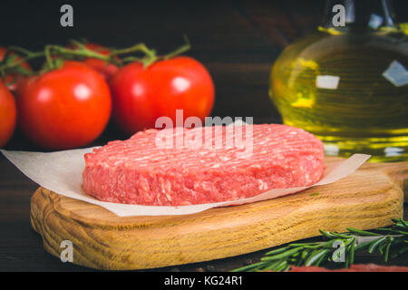 Raw Burger auf Pergamentpapier mit Rosmarin, Öl und Tomaten. Holz- braunen Hintergrund Stockfoto