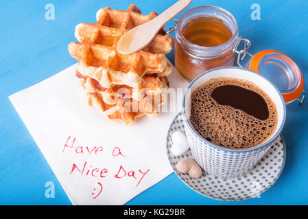 Tasse Kaffee mit Zucker in Herzform, selbstgemachte Waffeln mit Honig und eine Serviette mit schönen Tag noch Nachricht, auf blauem Holztisch. Stockfoto