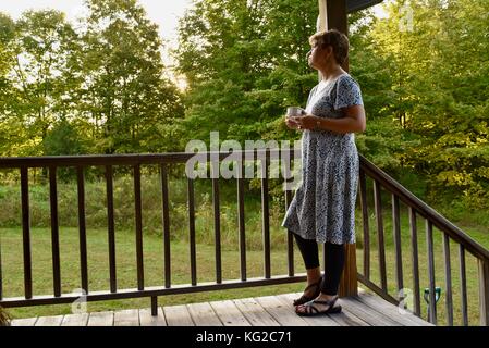 Frau mittleren Alters in Kleid auf Kabine Veranda mit Tasse Kaffee bei Sonnenaufgang. Private, rustikalen Hütte durch dichten Wald von Bäumen umgeben ist. Stockfoto