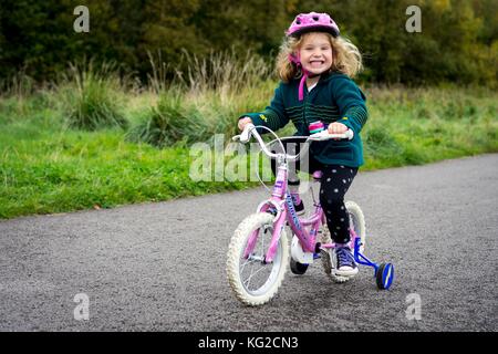 Junge, glückliche weibliches Kind reitet ihr rosa Fahrrad auf einem Pfad in den Park. Rimrose Valley Park, Sefton, Merseyside. Stockfoto