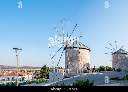 Hochauflösender Panoramablick auf Windmühlen, wo Menschen an einem sonnigen Tag in Alacati, einem beliebten Reiseziel, in einem Café in der Nähe essen und trinken Stockfoto