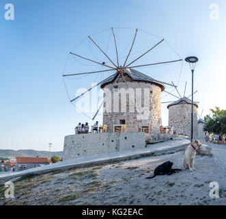 Hochauflösender Panoramablick auf Windmühlen, wo Menschen an einem sonnigen Tag in Alacati, einem beliebten Reiseziel, in einem Café in der Nähe essen und trinken Stockfoto