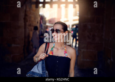 Attraktive trendy Frau Sonnenbrille tragen und mit einem großen Handtasche über die Schulter Einkaufen in der Stadt an der Seite mit einem Lächeln suchen Stockfoto