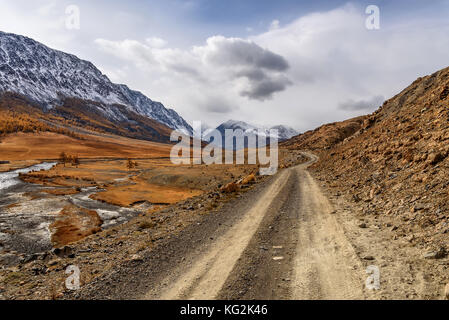 Die malerische Herbstlandschaft mit einer steinigen Piste, gewundenen Fluss, die Berge mit Schnee und goldenen Bäumen auf einem Hintergrund von Himmel und Cl abgedeckt Stockfoto