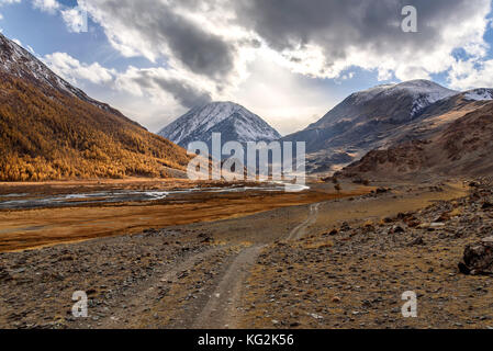 Die malerische Herbstlandschaft mit einer steinigen Piste, gewundenen Fluss, die Berge mit Schnee und goldenen Bäumen auf einem Hintergrund von Himmel und Cl abgedeckt Stockfoto