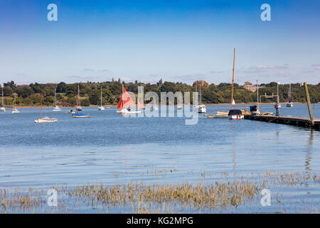 Eine kleine, traditionelle Yacht mit rotem Segelboot, segelt den Orwell hinauf, vorbei an vertäuten Booten in Pin Mill, Chelmondiston, Suffolk, Großbritannien Stockfoto