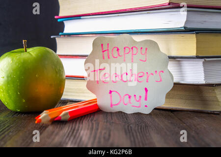 Konzept der Lehrer Tag. Objekte auf einer Schiefertafel Hintergrund. Bücher, grüner Apfel, Plakette: happy Lehrer Tag, Bleistifte und Kugelschreiber in einem Glas, Zweig mit Stockfoto