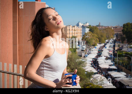 Jungen asiatischen Teenager Frau auf dem Balkon hoch geschlossenen Augen Sonnenlicht Blau Becher in die Hände städtischer Hintergrund Stadt Lebensstil Stockfoto