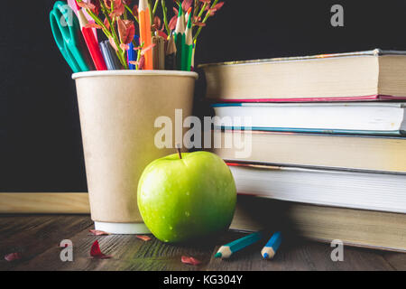 Konzept der Lehrer Tag. Objekte auf einer Schiefertafel Hintergrund. Bücher, grüner Apfel, Bleistifte und Kugelschreiber in einem Glas, Zweig, mit Blätter im Herbst. Stockfoto