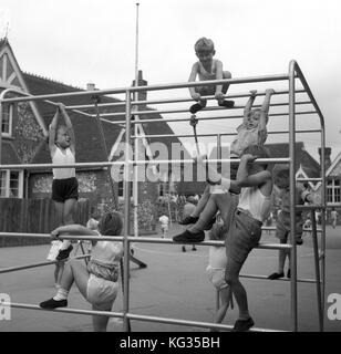 In den 1950er Jahren spielen historische Jungen und Mädchen aus dem Kindergarten auf einem Metallkletterrahmen draußen in ihrer Dorfschule auf dem Spielplatz und machen eine PE-Klasse, einige davon in ihrem Gym-Kit, England, Großbritannien. Stockfoto