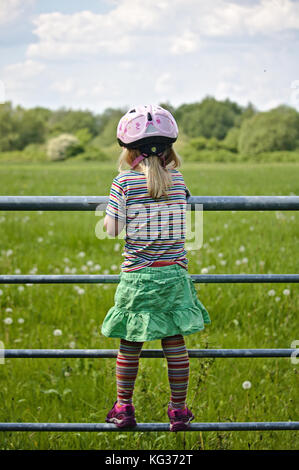 Kleines Mädchen tragen ein gestreiftes T-Shirt, Grün Rock und ein rosa Fahrrad Helm, stehend auf einem Feld Gate mit Blick auf ein grünes Feld voller Löwenzahn Stockfoto