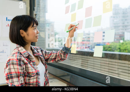 Ziemlich attraktive Frau vor der Glaswand ständige Arbeitsgruppe und mit Farbstift schreiben Hinweis auf Haftnotizen post-Papier. Stockfoto