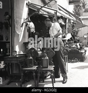 1960s, historisch, stöbert ein männlicher Tourist mit Kamera auf dem berühmten Flohmarkt Les Puces in Paris in einem Stall voller Möbel, Haushaltswaren und anderer Antiquitäten. Stockfoto