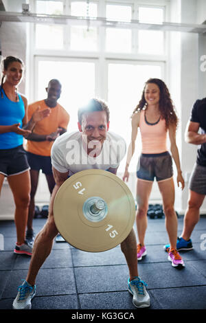 Lächelnden jungen Mann belasten Gewichte in einem Fitnessraum mit einer vielfältigen Gruppe von Freunde ihn Anfeuern im Hintergrund zu heben Stockfoto