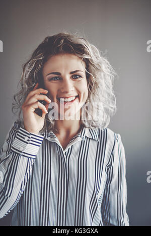 Porträt eines lächelnden jungen Geschäftsfrau mit blonden Haaren Gespräche am Mobiltelefon, während allein in einem Büro Stockfoto