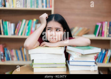 Asiatische Studentin langweilig lesen Buch an der Bibliothek mit einer Menge Bücher in der Universität. asiatische Student entmutigt lesen Buch für die Prüfung. Stockfoto