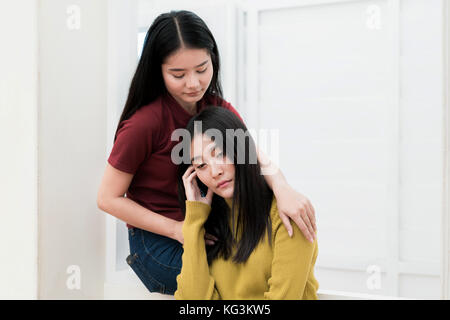 Schöne asiatische Frau umarmen depressiven Freund beim Sitzen auf Fenster zu Hause Stockfoto