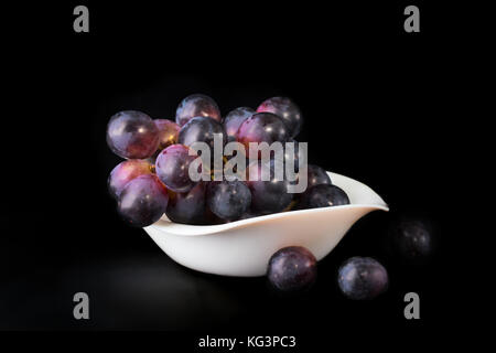 Cluster von schwarzen Trauben auf einem schwarzen Hintergrund. Trauben in eine weiße Platte liegen, einige Beeren liegen in der Nähe Stockfoto