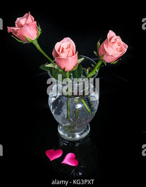 Rosa Rosen in einer Glasvase und zwei Herzen auf Schwarz. Drei Rosen und zwei Herzen von rosa Stoff auf schwarzem Hintergrund Stockfoto