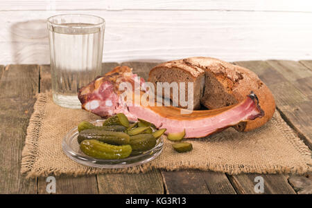 Produkte auf einem alten Holztisch, ländlichen Stil. Auf Serviette von einer Entlassung Brot, Schinken auf Kanten, Essiggurken und ein Messer, in der Nähe von einem Glas Wasser liegt. Stockfoto