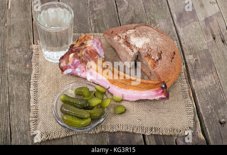 Produkte auf einem alten Holztisch, ländlichen Stil. Auf Serviette von einer Entlassung Brot, Schinken auf Kanten, Essiggurken und ein Messer, in der Nähe von einem Glas Wasser liegt. Stockfoto