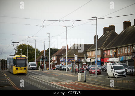 Eine gelbe Manchester Metrolink tram an Hollyhedge Straße, Benchill, Wythenshawe. Vorbei eine Parade von unabhängigen lokalen Geschäften Stockfoto