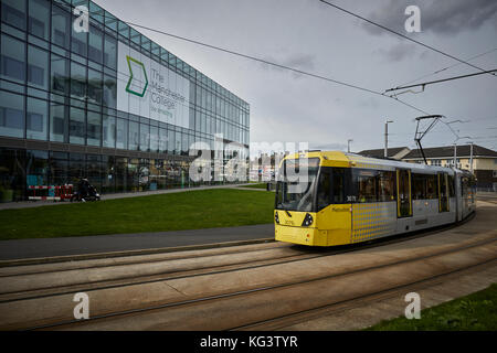 Eine gelbe Manchester Metrolink tram an Hollyhedge Straße, Benchill, Wythenshawe, Manchester College moderne geruhte Gebäude Stockfoto