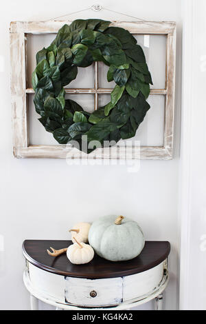 Altes Bauernhaus Fenster mit einem hausgemachten magnolia Blatt Kranz an einer Innenwand über rustikale half moon Tabelle mit erbstück Kürbisse aufgehängt eingerichtet. Stockfoto