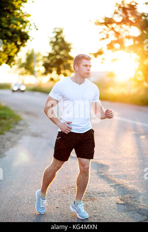Ein Athlet in einem weißen T-Shirt und Shorts Läufer in Abend, joggen. Ein Mann im Park hört, ein Hörbuch, ein zuversichtlich. Sommer Lifestyle, mo Stockfoto