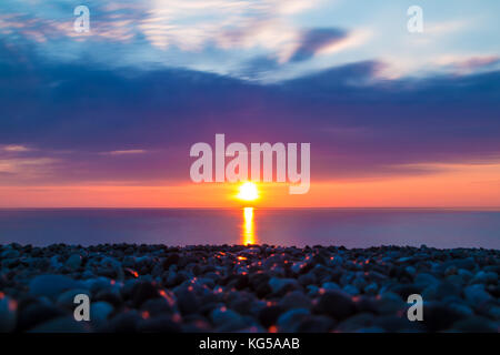 Lange Belichtung auf den Kiesstrand vor dem Hintergrund der untergehenden Sonne über dem Meer Stockfoto