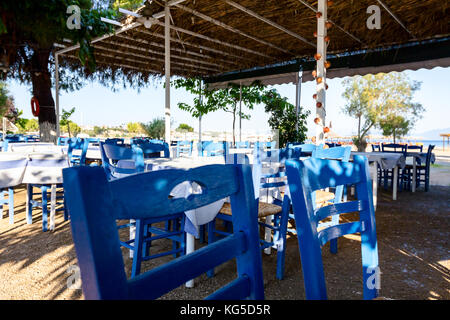 Stühle und Tische in typische griechische Taverne in der Morgensonne mit Schatten am Meeresufer. Stockfoto