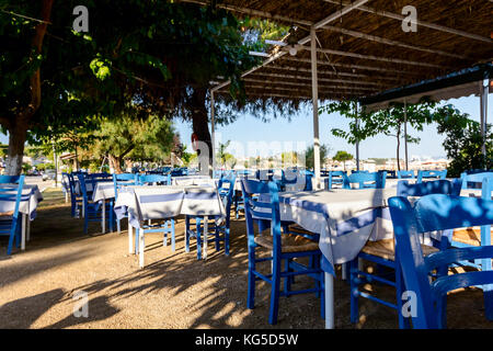 Stühle und Tische in typische griechische Taverne in der Morgensonne mit Schatten am Meeresufer. Stockfoto