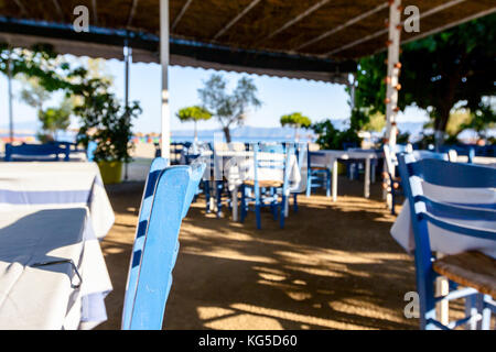 Stühle und Tische in typische griechische Taverne in der Morgensonne mit Schatten am Meeresufer. Stockfoto