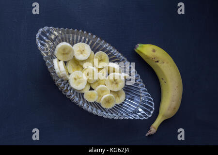 Bananenscheiben in Glasschale mit ganze ungeschälte Banane auf schwarzem Hintergrund - Ansicht von oben Foto Stockfoto