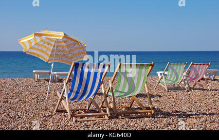 Eine Reihe von leeren Liegestühlen unter einem blauen Himmel auf einem Kieselstrand mit einem Wetter gebeizt Sonnenschirm von der Professur an der Kamera am nächsten ist Stockfoto