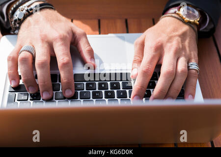 Anonyme männliche Unternehmer oder Arbeitnehmer mit Armbänder, Ringe, Uhren und Zubehör auf Hände und Finger an einer hölzernen orange Tisch sitzen und tippen auf Stockfoto
