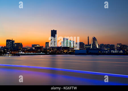 Yokohama, Japan die Skyline in der Dämmerung mit die Lichter der Stadt und orange sky als Boot Verkehr schafft helle Streifen auf dem Wasser und den Berg Fuji zwischen Gebäuden Stockfoto