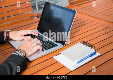 Anonyme männliche Unternehmer oder Arbeitnehmer im schwarzen Anzug, Armbänder und Zubehör auf Händen draußen sitzen auf einem hölzernen orange Tisch und Tippen und Arbeiten Stockfoto