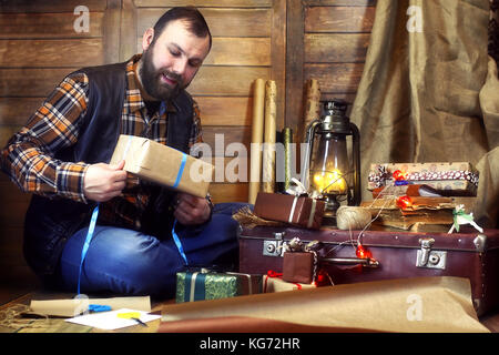 Bärtiger Mann im Hemd und Weste packs Weihnachten Geschenke an die Kamera in eine hölzerne Dekorationen posing Stockfoto