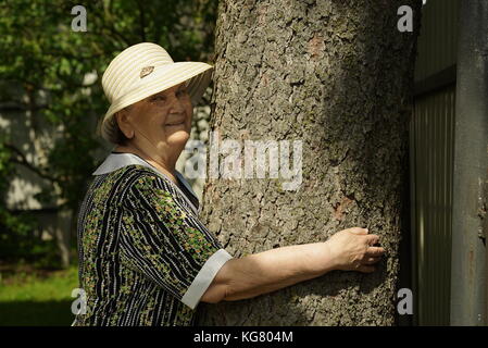 Ältere, reife Frau 80 s in White hat umarmt Baum ihre Hände in den Wald gekleidet Jahren. Frau ist Energie der Natur erhoben. Entspannung in einem Summ Stockfoto