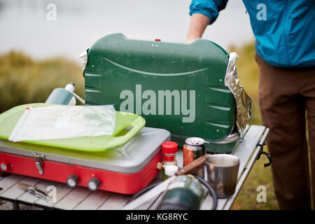 Mann, ein Camping Kocher Gas im Freien auf einen kleinen Tisch mit Zutaten und Gewürze, wie er sich vorbereitet, um eine Mahlzeit zu kochen Stockfoto