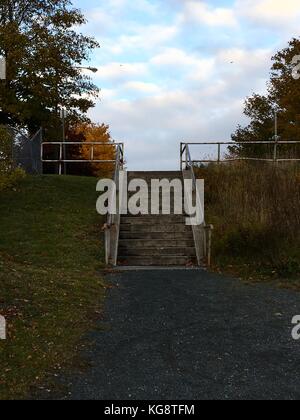 Beton Treppe, die aus einer Kiesgrube Wanderweg zu einer Stadt Straße. blauer Himmel mit Wolken Licht am Ende der Treppe sichtbar, Bäume im Herbst Farben Stockfoto