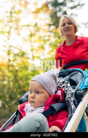 Ausführen von Mutter mit kontemplativen Kind im Kinderwagen bei Mutterschaft im Herbst Sonnenuntergang und Wald landschaft. Joggen oder Walken Frau Stockfoto