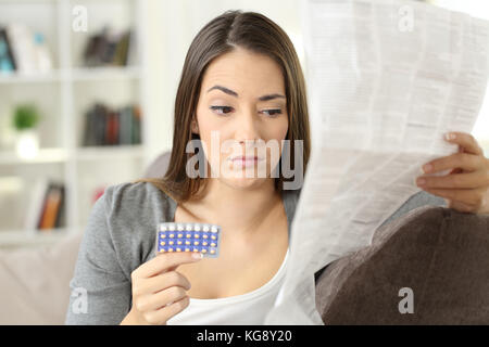 Verdächtige Frau liest eine Broschüre nach der Einnahme von empfängnisverhütenden Pillen sitzen auf einer Couch im Wohnzimmer in einem Haus innen Stockfoto