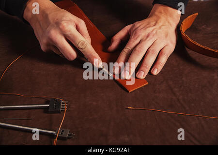 Eine Nahaufnahme eines Skinner Mann macht ein echtes Leder Gürtel, schneidet die Haut mit einem speziellen Messer auf den Tisch der Rinde, eine Sicht von oben Stockfoto