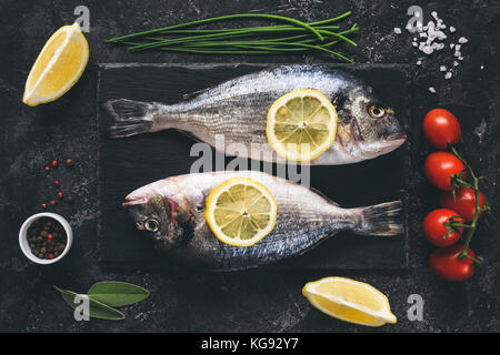 Frische Seeluft Strahl oder dorado Fisch-, Zitronen-, Kräuter, Gewürze und Gemüse auf schwarzen Stein Schiefer Hintergrund. Ansicht von oben. Frischer Fisch bereit für das Kochen. Getönten imag Stockfoto